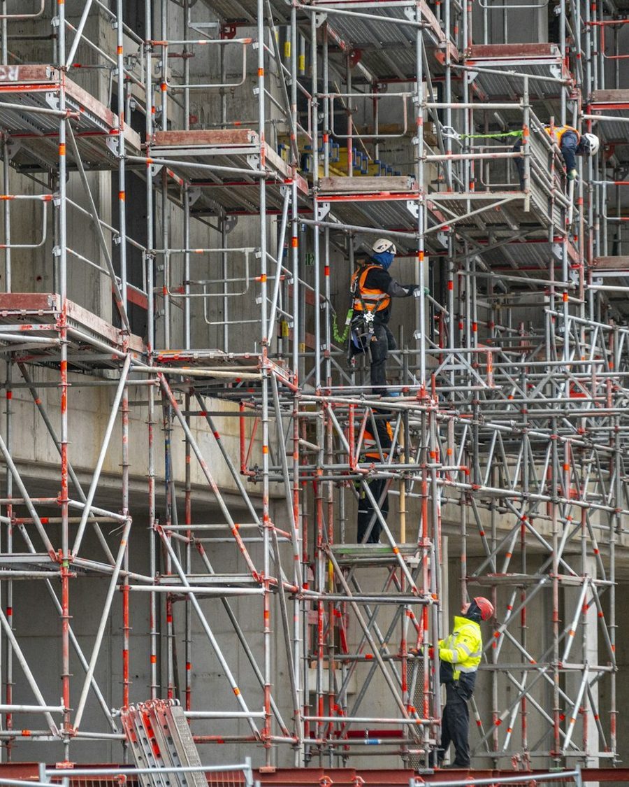 Gerüst mit mehreren Ebenen vor einer Hausfassade, zwei Arbeiter mit Helm und Warnweste auf dem Gerüst